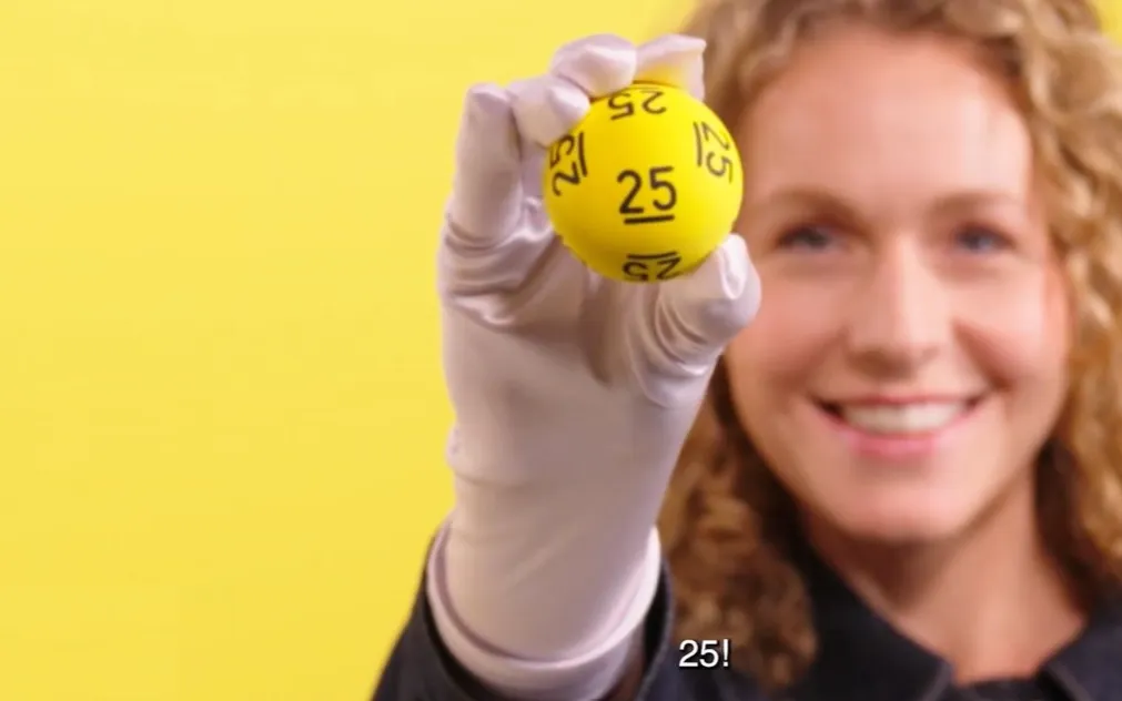 Een hand met een gele bal waarop het nummer 25 staat, tegen een gele achtergrond.