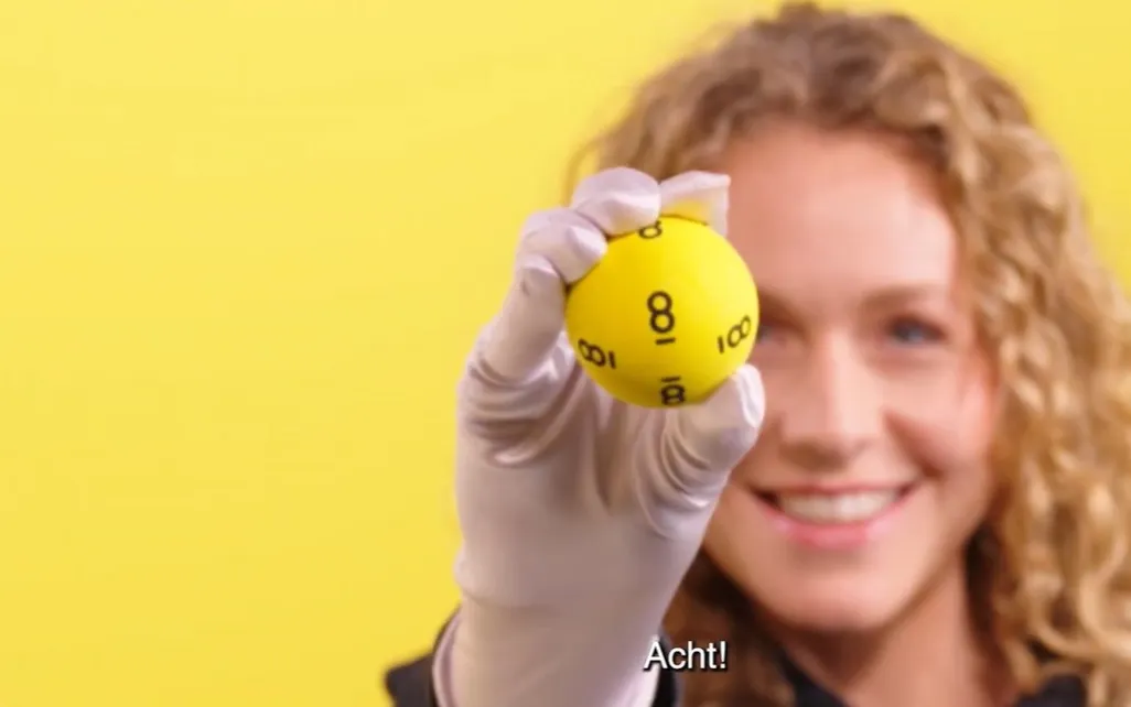 Een persoon in een witte handschoen houdt een gele bal omhoog met het nummer acht, tegen een gele achtergrond.