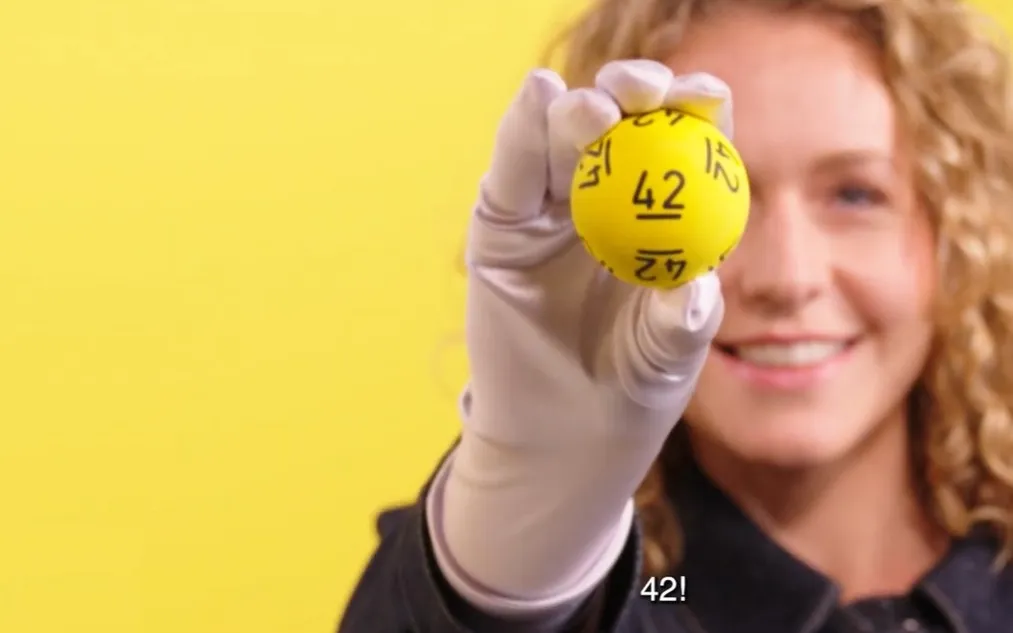 Persoon houdt een gele Jackpotbal met het nummer 42 omhoog tegen een gele achtergrond.
