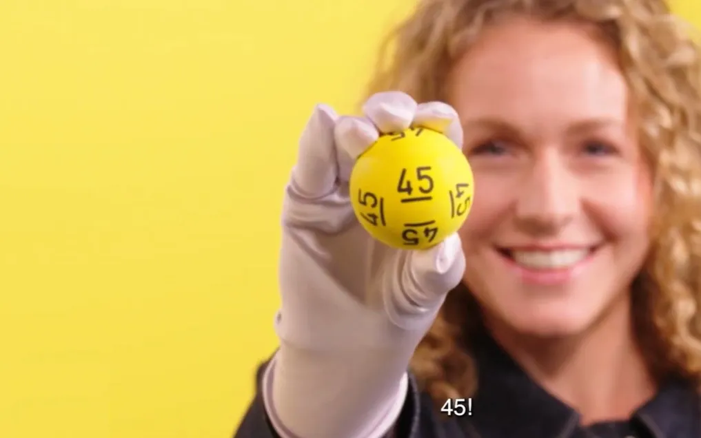 Een hand houdt een gele Lotto-bal met het nummer 45 omhoog, tegen een gele achtergrond.