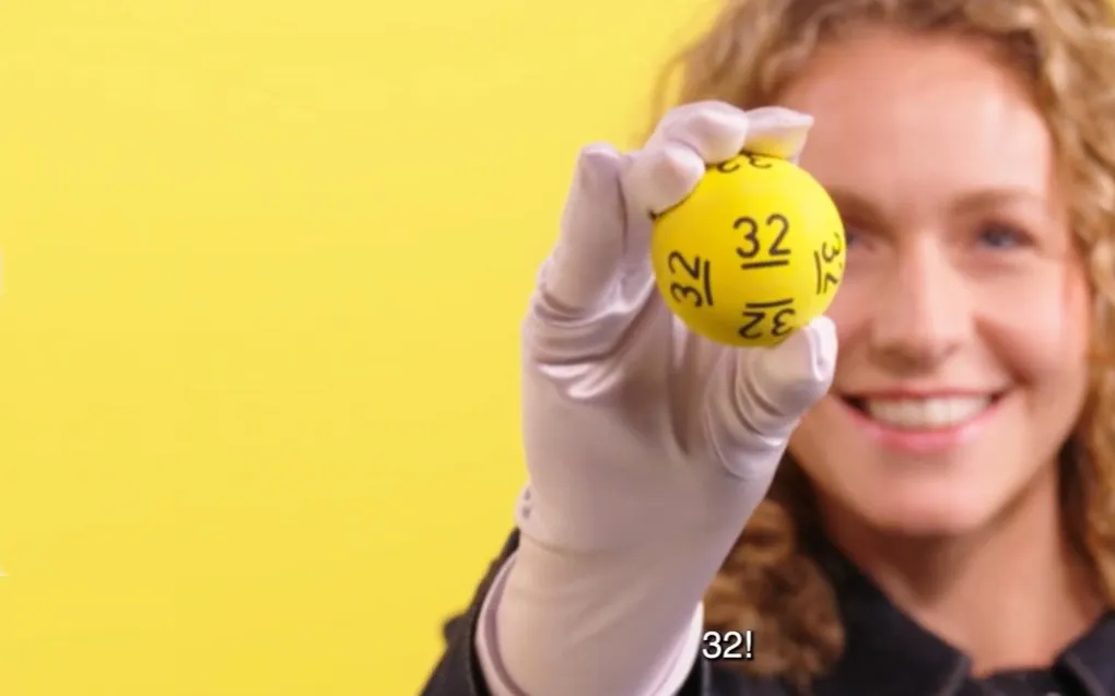Een persoon met een witte handschoen houdt een gele bal met het nummer 32 omhoog, op een gele achtergrond.