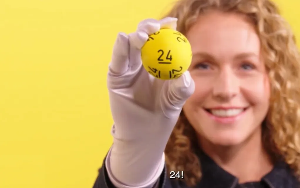 Een hand in een witte handschoen houdt een gele bal met het nummer 24 omhoog tegen een gele achtergrond.