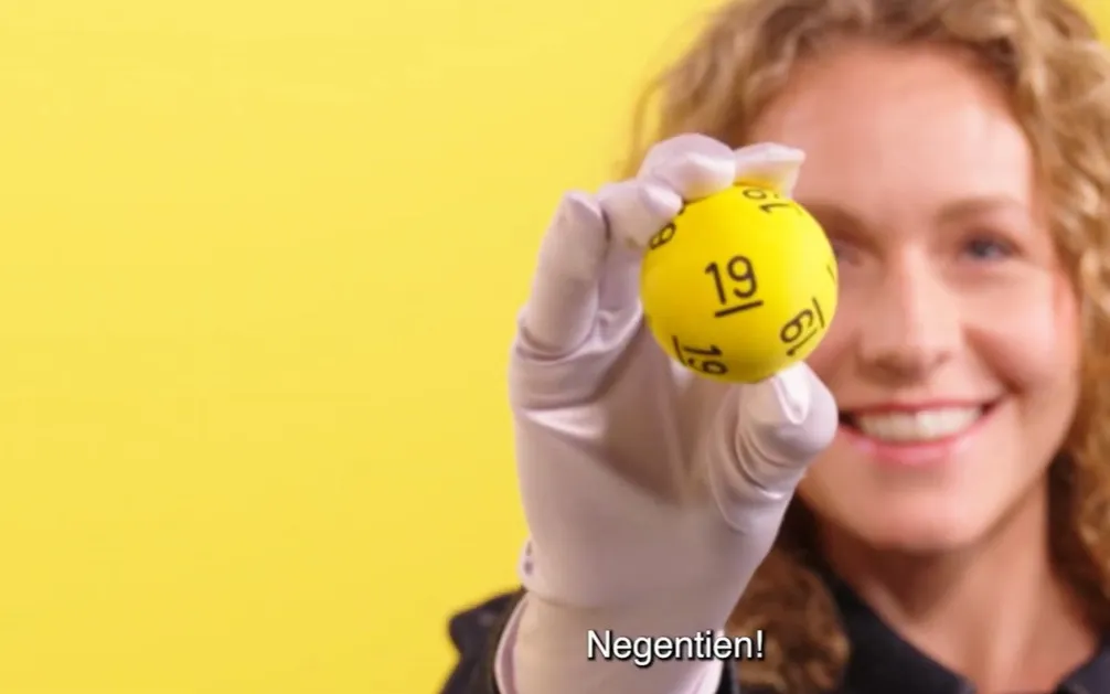 Een persoon houdt een gele bal met een nummer omhoog tegen een gele achtergrond.