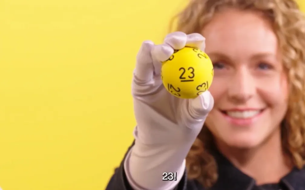 Hand houdt een gele bal met het nummer 23 omhoog tegen een gele achtergrond.