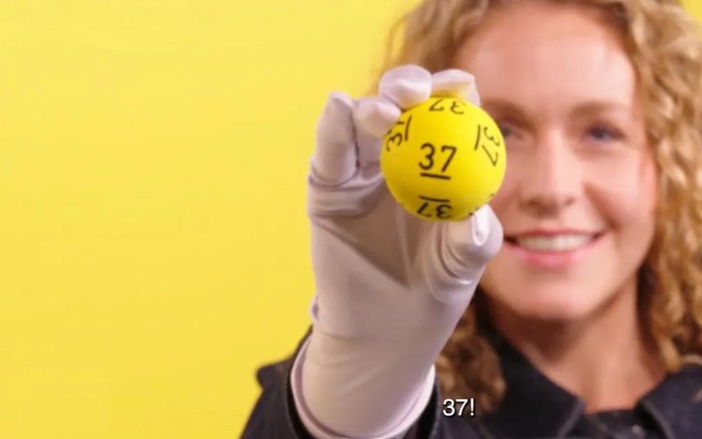 Een persoon houdt een gele Jackpotbal met het nummer 37 omhoog tegen een gele achtergrond.