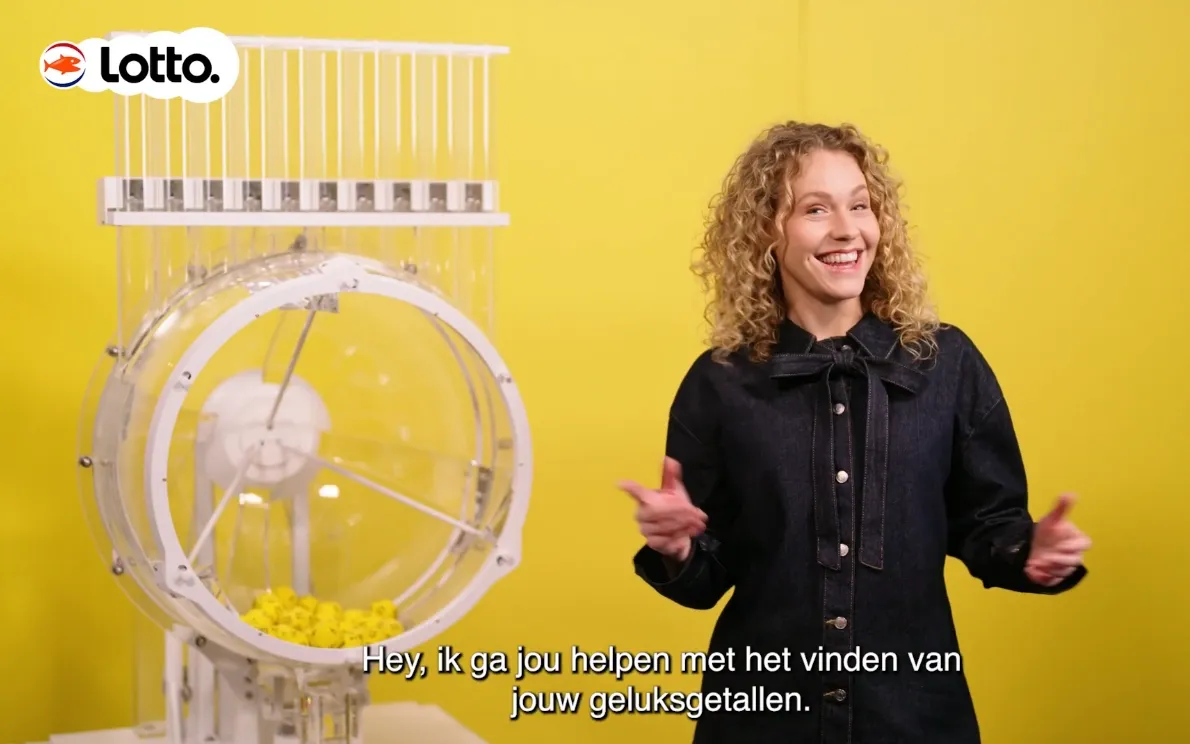 Presentator staat naast een Lotto Jackpotmachine op een gele achtergrond.