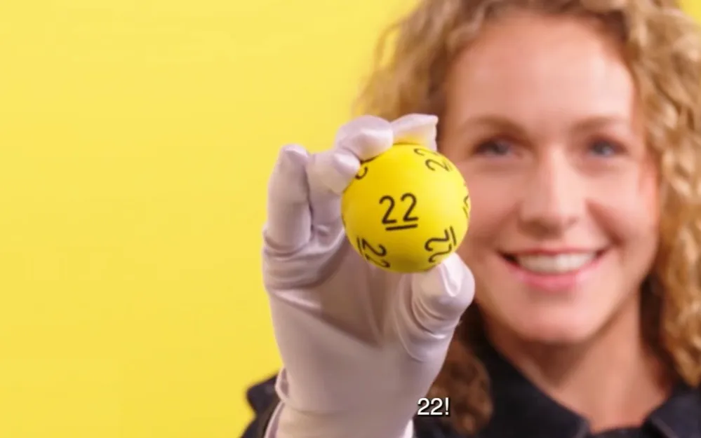 Een persoon houdt een gele loterijbal met het nummer 22 omhoog.