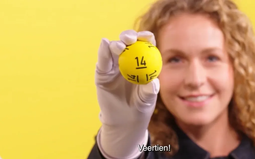 Een gele bal met het nummer 14, vastgehouden door een persoon met handschoenen, tegen een gele achtergrond.