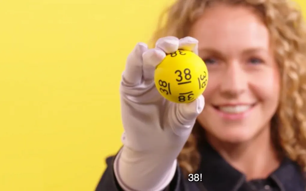 Een hand die een gele bal met het nummer 38 omhooghoudt, tegen een gele achtergrond.