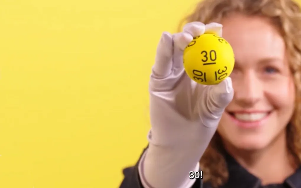 Een persoon houdt een gele Lotto-bal met het nummer 30 omhoog tegen een gele achtergrond.