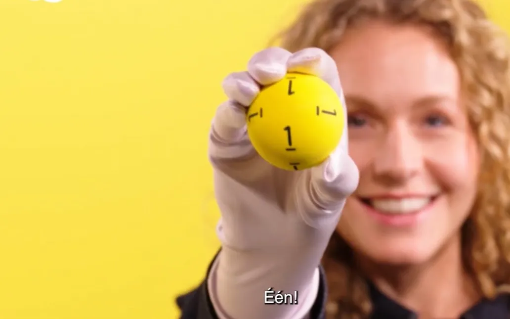 Een persoon houdt een gele bal met het getal 1 omhoog tegen een gele achtergrond.