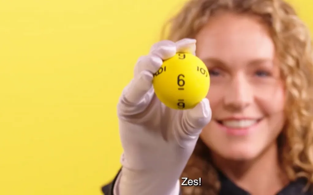 Persoon die een gele Lotto-bal met het nummer 6 omhooghoudt voor een gele achtergrond.