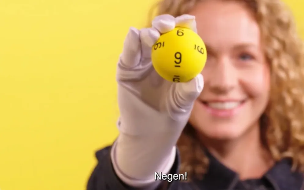 Een persoon houdt een gele bal omhoog met het nummer negen op een gele achtergrond.