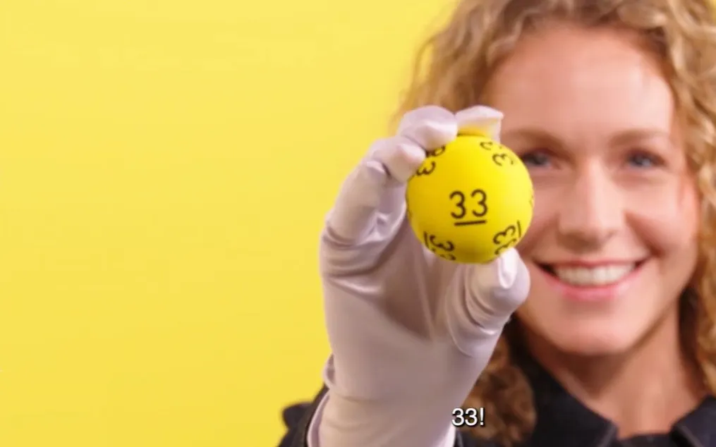 Een hand houdt een gele ballon met het nummer 33 omhoog tegen een gele achtergrond.
