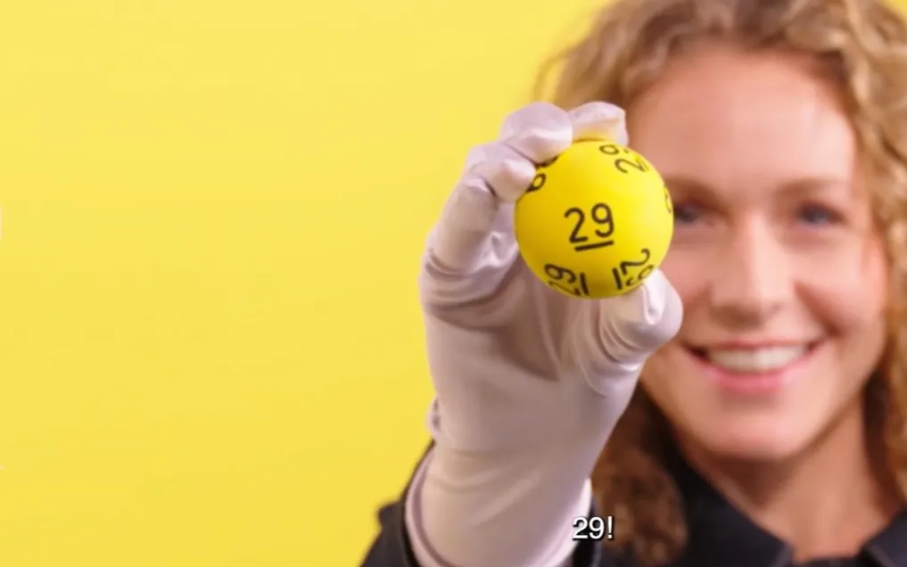 Een persoon houdt een gele bal met het nummer 29 omhoog, tegen een gele achtergrond.