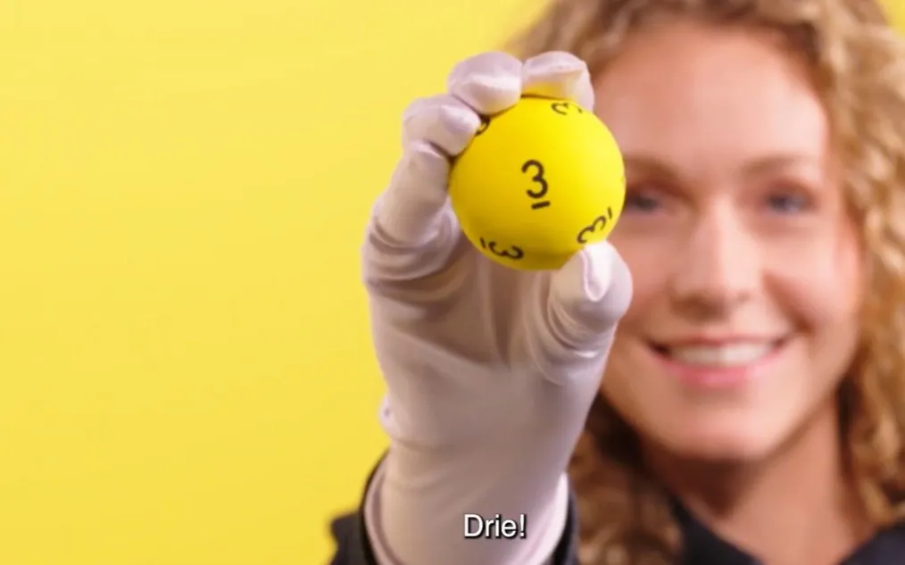 Een hand houdt een gele bal met het nummer 3 omhoog tegen een gele achtergrond.