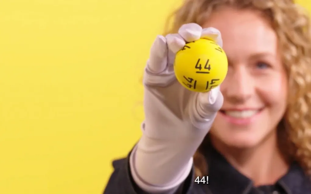 Een persoon houdt een gele bal met het nummer 44 omhoog, tegen een gele achtergrond.