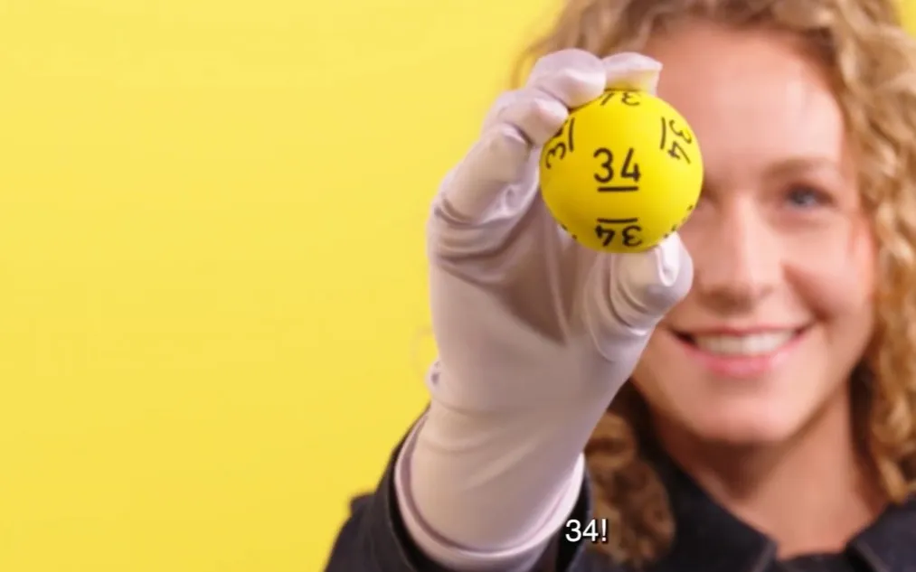 Een gelukkige lotto-omroepster toont een gele bal met het nummer 34 op een gele achtergrond.