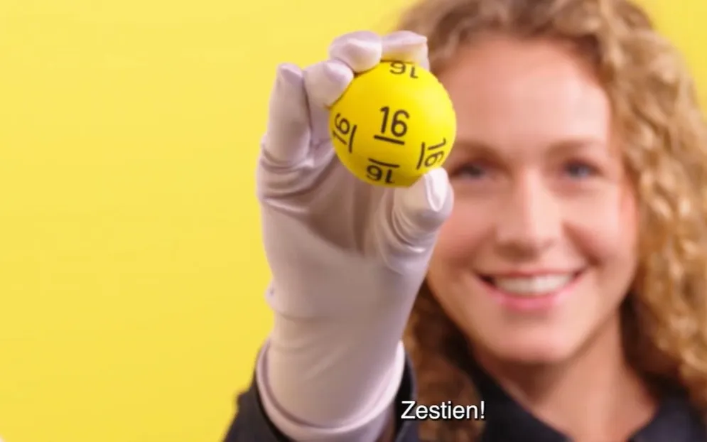 Een hand houdt een gele bal met het nummer 16 omhoog tegen een gele achtergrond.